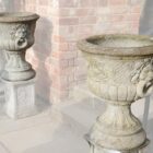A pair of reclaimed decorative lions head urns in composite stone, showing a fine patina of discolouration and lichen patches, ready for planting