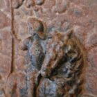 A close up of an antique and weathered cast iron fireback showing a regal horse mounted figure within an arch of grape and vine motif.