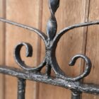 A detail of decorative scrollwork on a reclaimed wrought iron gate with peeling black paint revealing patches of surface corrosion