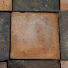 A close up of one of the red tiles from a batch of reclaimed red and blue quarry tiles laid in a classic chequerboard pattern, showing the pitted surfaces and discolouration commensurate with age