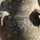 Detail of a well weathered composite stone vase with four handles and a slim profile showing a lovely patina of discolouration and lichen.